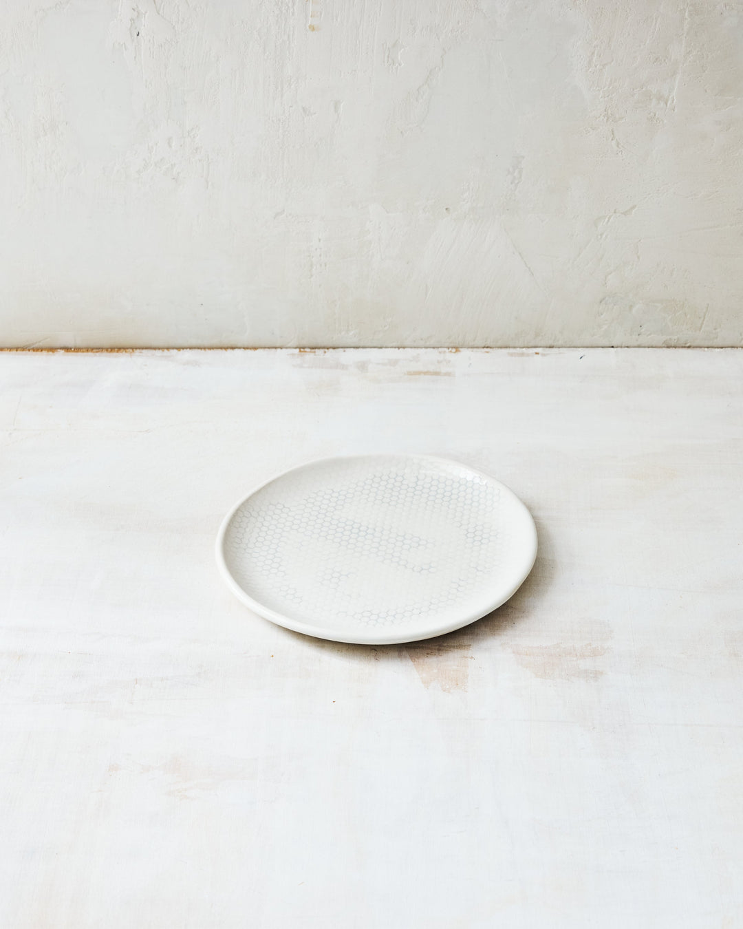 DBO HOME Honeycomb Bread Plate in handmade porcelain with a white gloss glaze and pale grey-inlaid honeycomb texture, photographed at a low angle on a whitewashed surface.