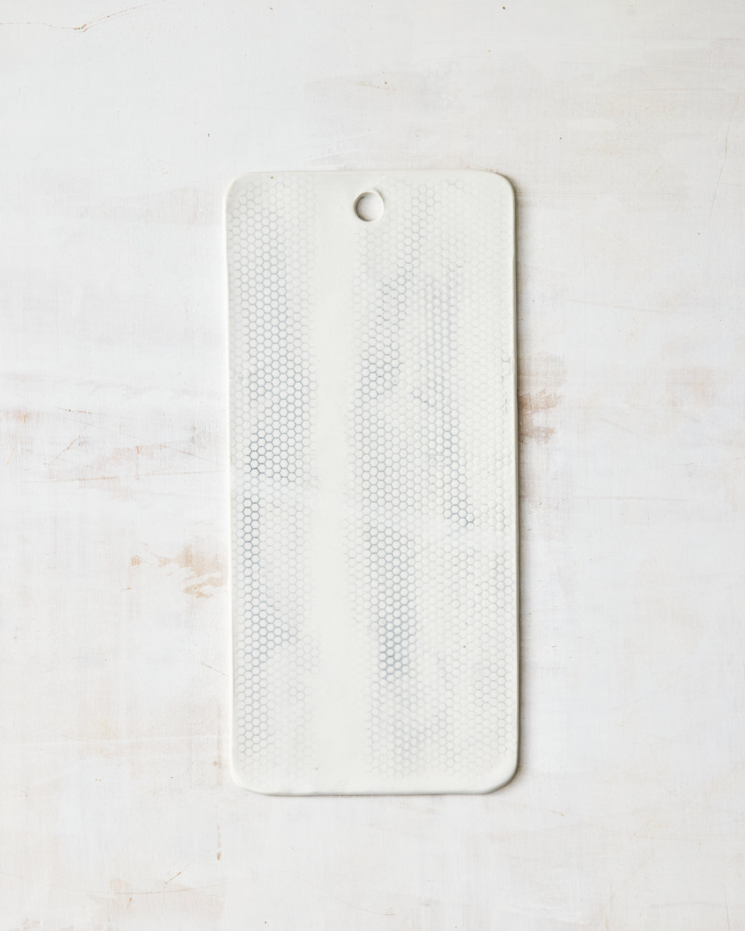 DBO HOME Honeycomb Charcuterie Board in handmade porcelain with a white gloss glaze and pale grey-inlaid honeycomb texture, narrow rectangular form with a hanging hole at the top, photographed overhead on a white painted wood surface.