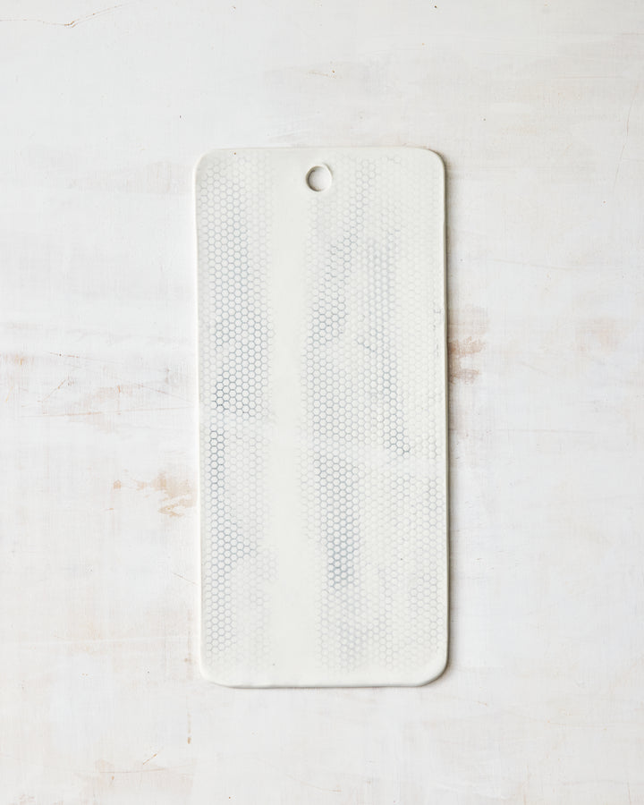 DBO HOME Honeycomb Charcuterie Board in handmade porcelain with a white gloss glaze and pale grey-inlaid honeycomb texture, narrow rectangular form with a hanging hole at the top, photographed overhead on a white painted wood surface.