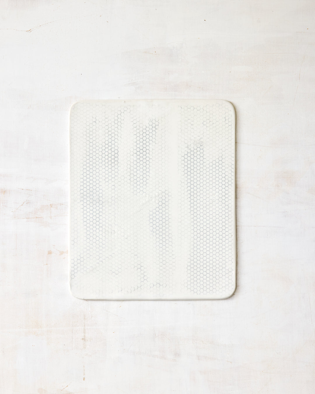 DBO HOME Honeycomb Cheese Board in handmade porcelain with a white gloss glaze and pale grey-inlaid honeycomb texture, wide square form with softly rounded corners, photographed overhead on a white painted wood surface.