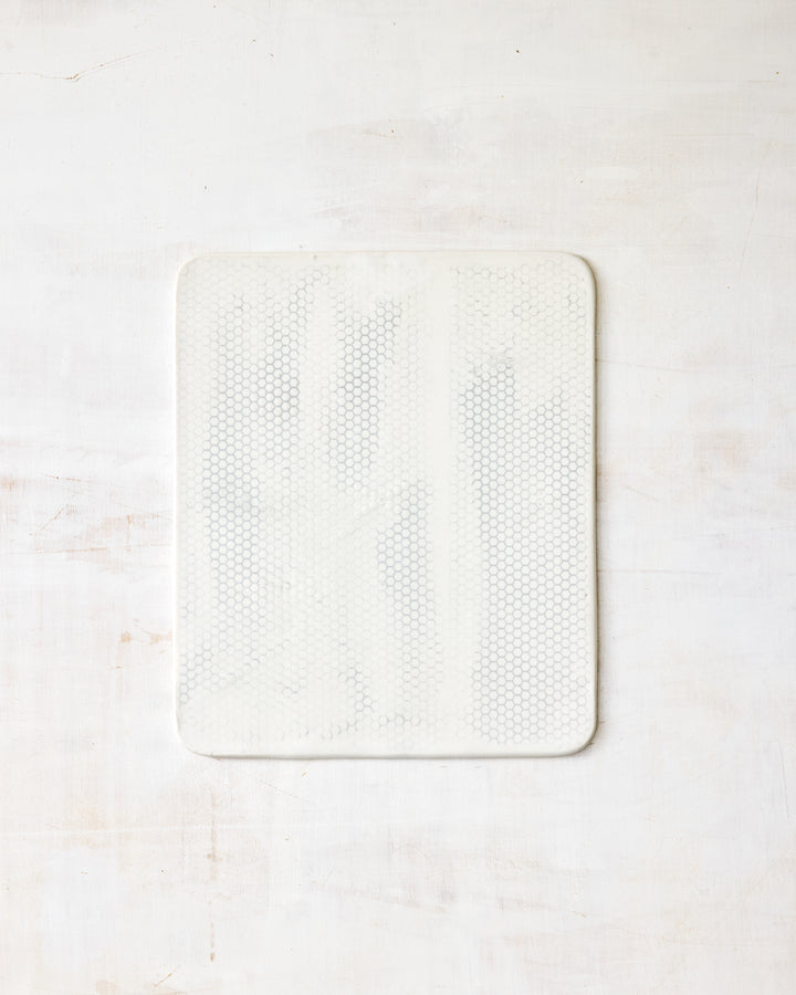 DBO HOME Honeycomb Cheese Board in handmade porcelain with a white gloss glaze and pale grey-inlaid honeycomb texture, wide square form with softly rounded corners, photographed overhead on a white painted wood surface.