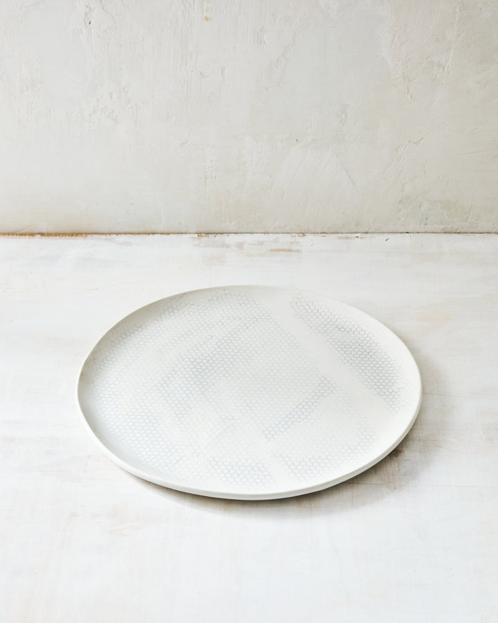 DBO HOME Honeycomb Dinner Plate in handmade porcelain with a white gloss glaze and pale grey-inlaid honeycomb texture, photographed at a low angle on a whitewashed surface.