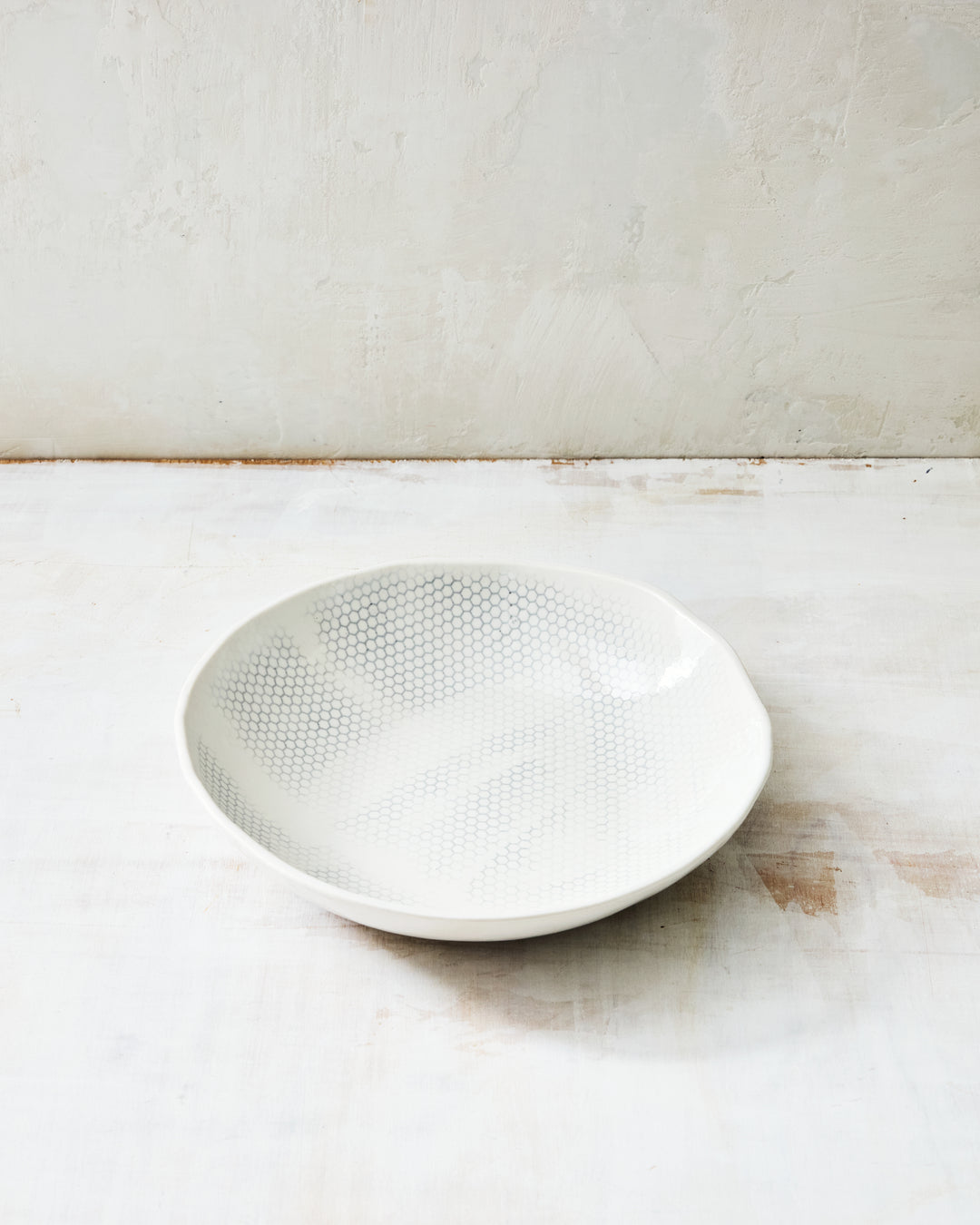 DBO HOME Honeycomb Everything Bowl in handmade porcelain with a white gloss glaze and pale grey-inlaid honeycomb texture, wide and shallow with an organic rim, photographed at a low angle on a whitewashed surface.