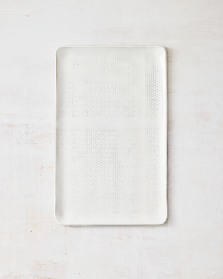 DBO HOME Honeycomb Large Platter in handmade porcelain with a white gloss glaze and pale grey-inlaid honeycomb texture, wide rectangular form with softly rounded corners, photographed overhead on a white painted wood surface.