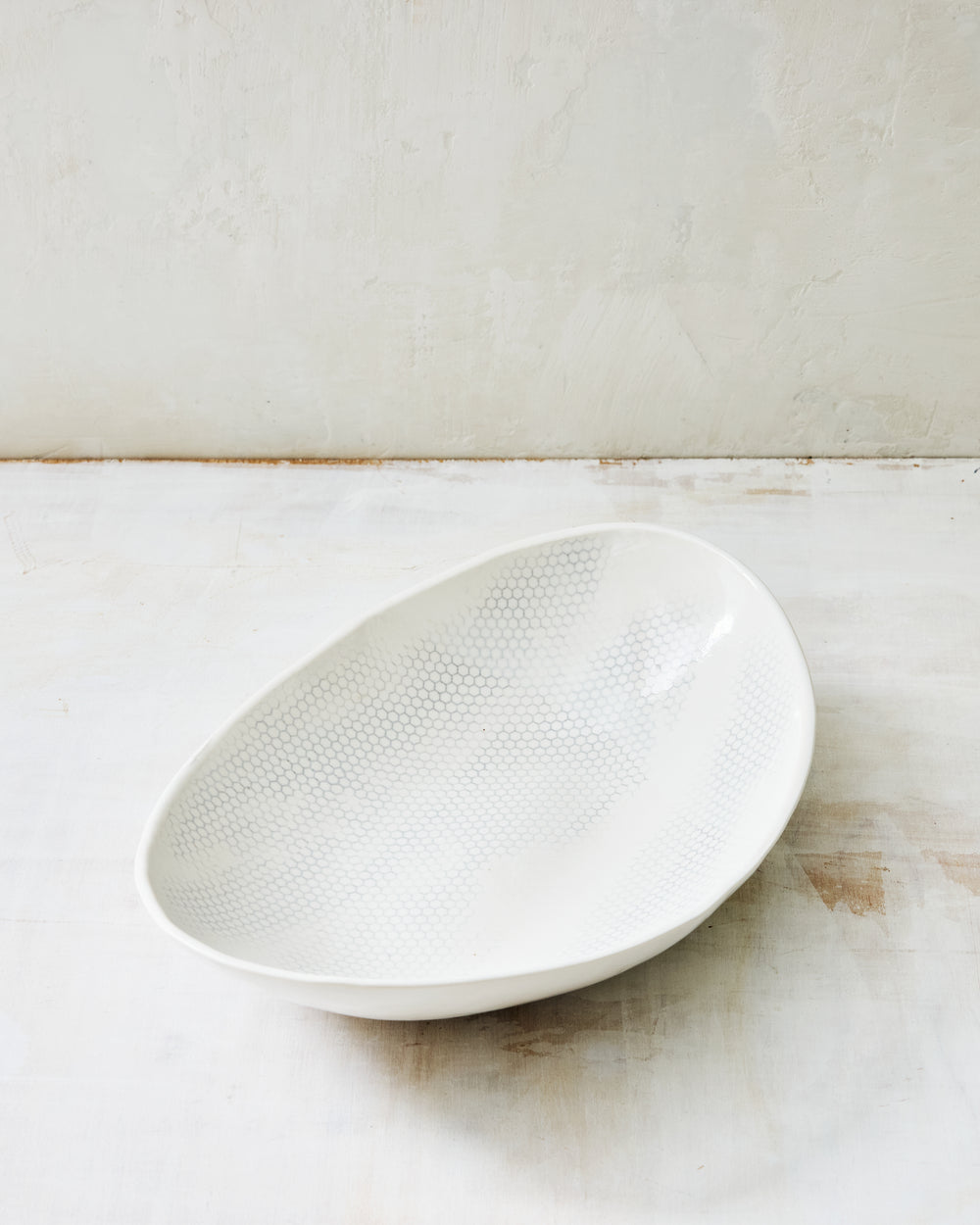 DBO HOME Honeycomb Oval Bowl in handmade porcelain with a white gloss glaze and pale grey-inlaid honeycomb texture, deep organic oval form with softly irregular edges, photographed at a low angle on a whitewashed surface.