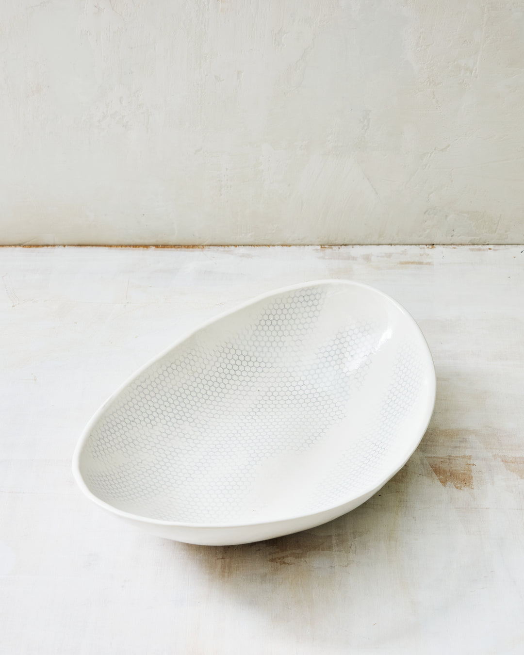 DBO HOME Honeycomb Oval Bowl in handmade porcelain with a white gloss glaze and pale grey-inlaid honeycomb texture, deep organic oval form with softly irregular edges, photographed at a low angle on a whitewashed surface.