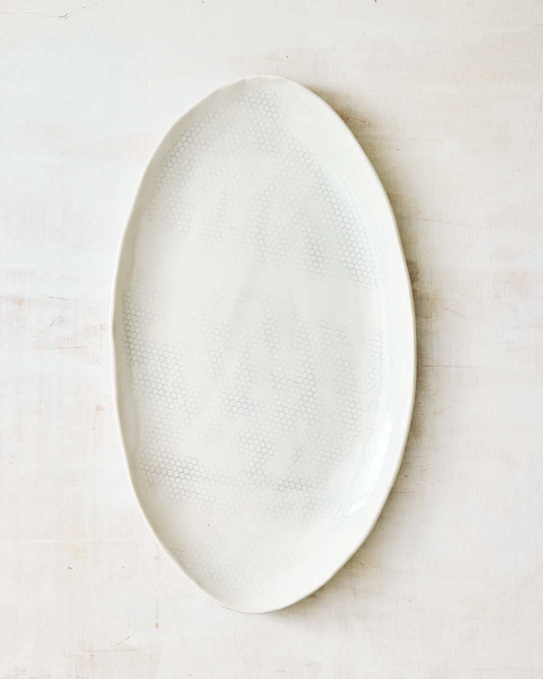 DBO HOME Honeycomb Small Oval Platter in handmade porcelain with a white gloss glaze and pale grey-inlaid honeycomb texture, organic oval form with a softly irregular rim, photographed overhead on a white painted wood surface.