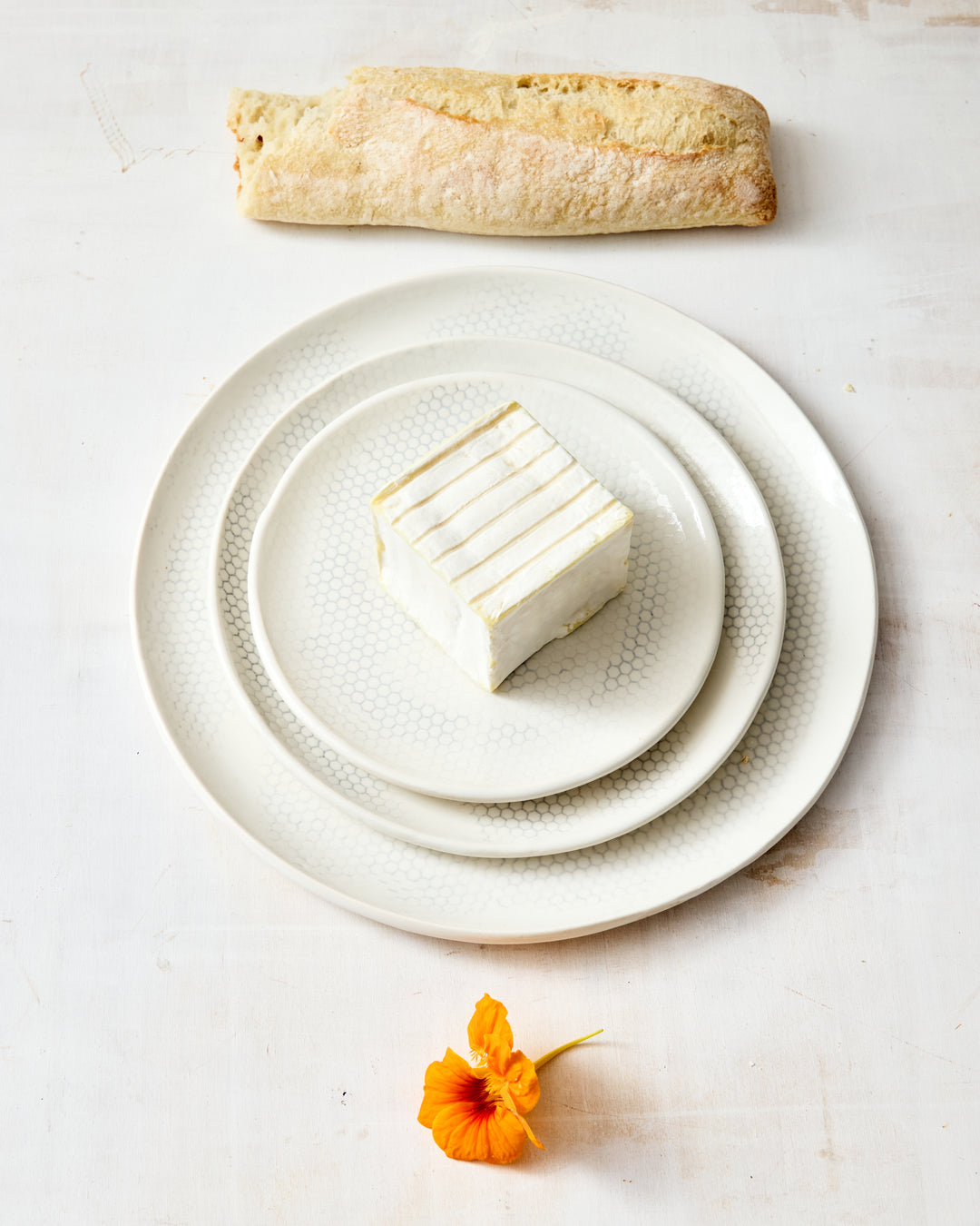 Three handmade porcelain plates from DBO HOME's Honeycomb collection stacked overhead — bread, salad, and dinner plate in a white gloss glaze with pale grey-inlaid honeycomb pattern, styled with cremini mushrooms on a whitewashed surface.