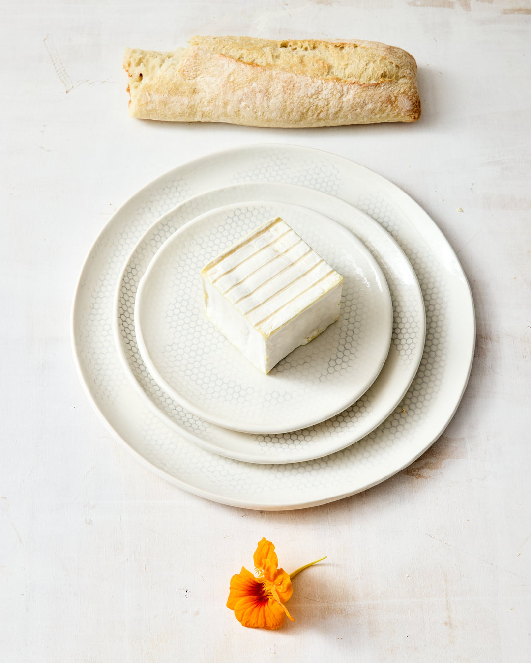Three handmade porcelain plates from DBO HOME's Honeycomb collection stacked overhead — a bread plate, salad plate, and dinner plate in a white gloss glaze with pale grey-inlaid honeycomb pattern, styled with cremini mushrooms on a whitewashed surface.