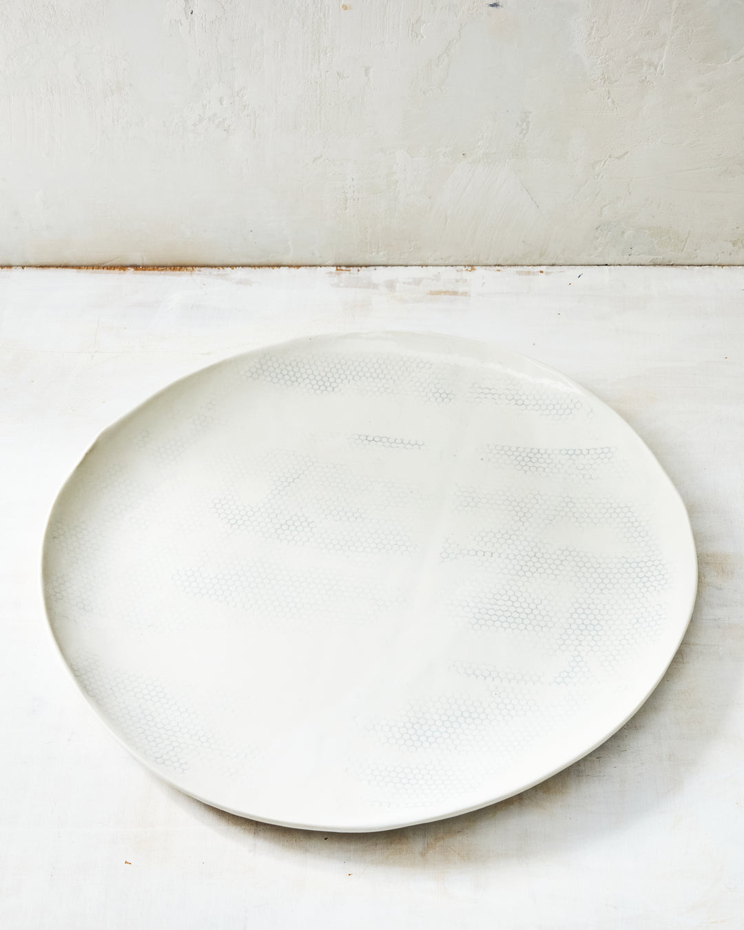 DBO HOME Honeycomb Round Tray in handmade porcelain with a white gloss glaze and pale grey-inlaid honeycomb texture, wide flat circular form with a low rim, photographed at a low angle on a whitewashed surface.
