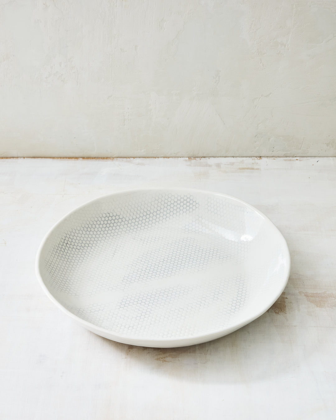 DBO HOME Honeycomb Serving Bowl in handmade porcelain with a white gloss glaze and pale grey-inlaid honeycomb texture, versatile wide form with gently curved sides, photographed at a low angle on a whitewashed surface.