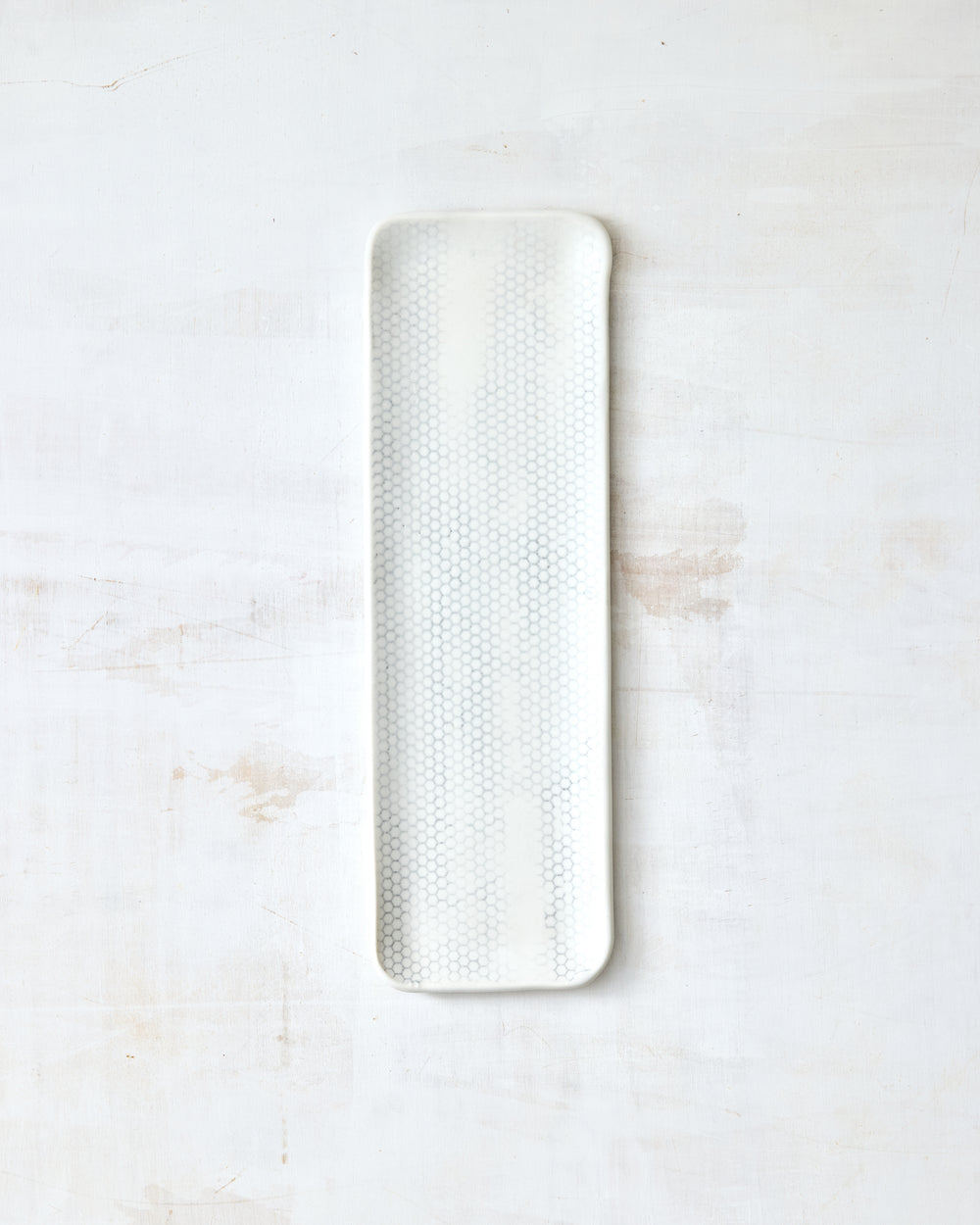 DBO HOME Honeycomb Tray in handmade porcelain with a white gloss glaze and pale grey-inlaid honeycomb texture, narrow elongated form with softly rounded corners, photographed overhead on a white painted wood surface.