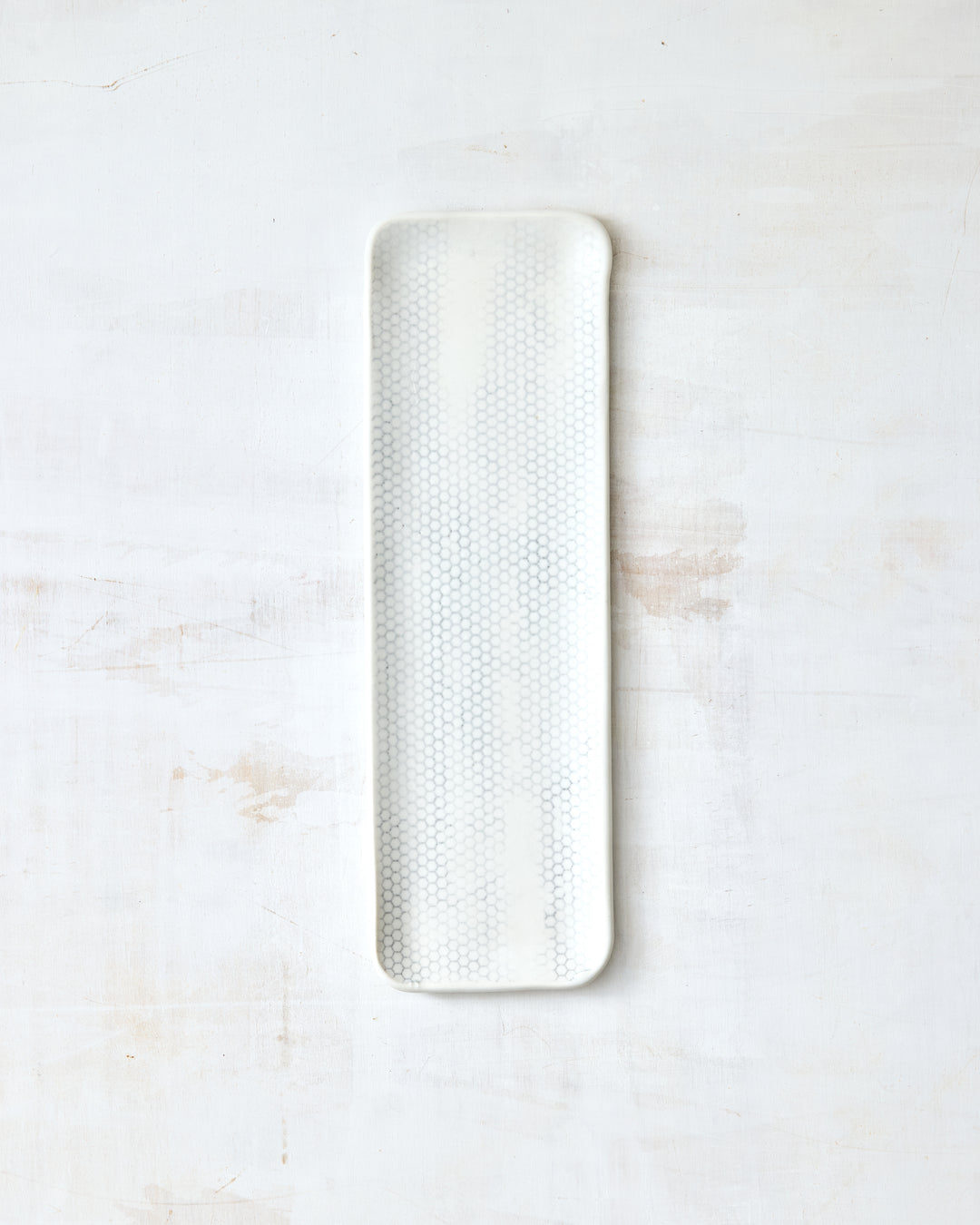 DBO HOME Honeycomb Tray in handmade porcelain with a white gloss glaze and pale grey-inlaid honeycomb texture, narrow elongated form with softly rounded corners, photographed overhead on a white painted wood surface.