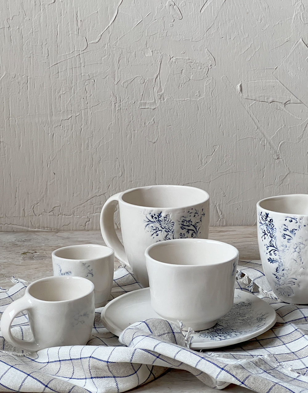 Kashmir ceramic mugs styled with coordinating handmade tableware, featuring blue floral patterns on white stoneware, arranged on a linen cloth for a casual, everyday table setting.
