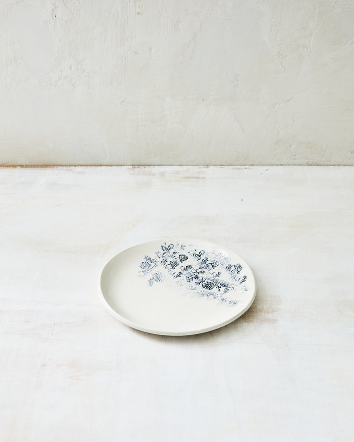 Small Kashmir bread plate in white porcelain featuring a delicate indigo blue floral pattern, photographed against a light, textured background.