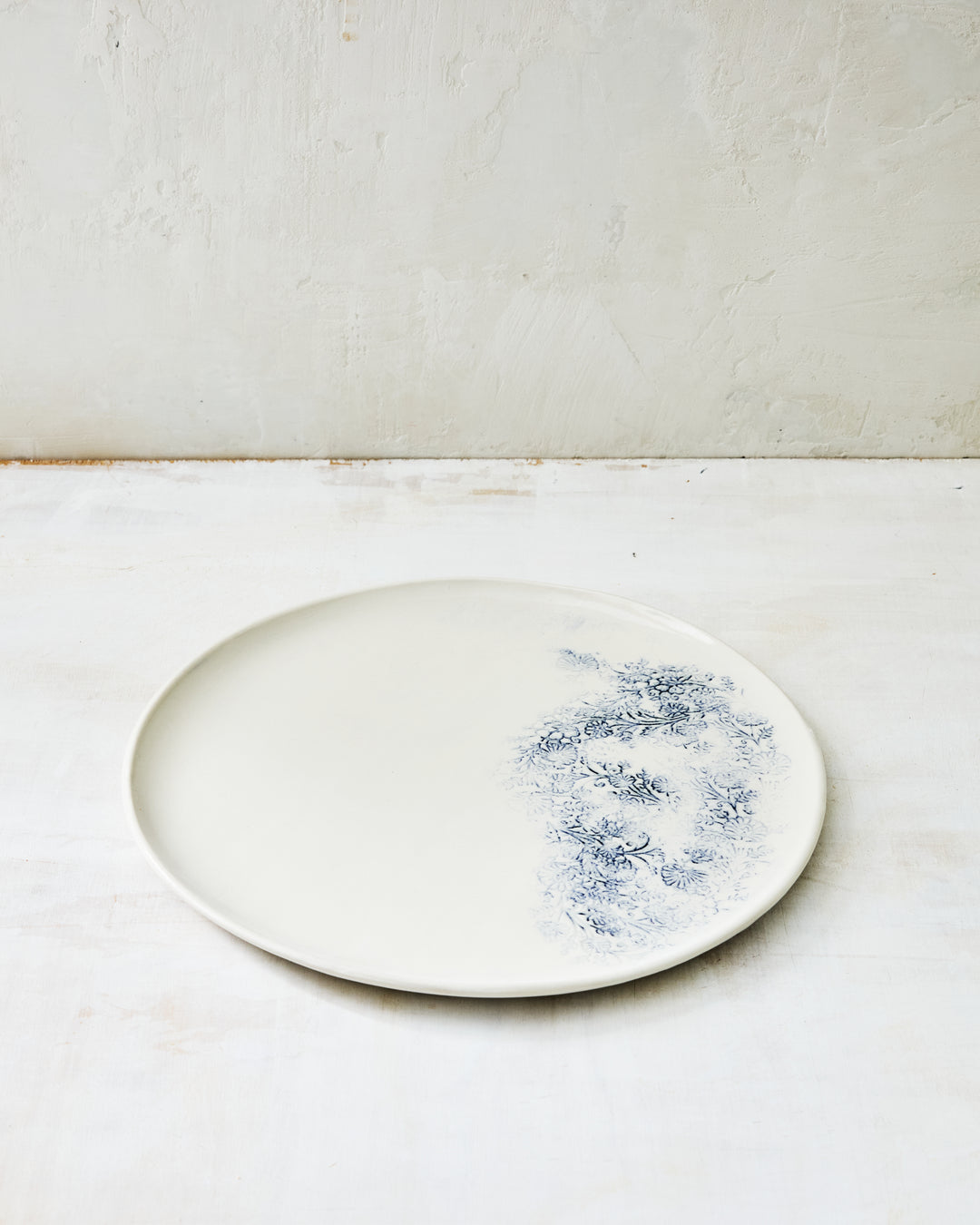 Handmade Kashmir dinner plate in white porcelain with a soft blue indigo floral motif along one edge, shown on a light textured surface.