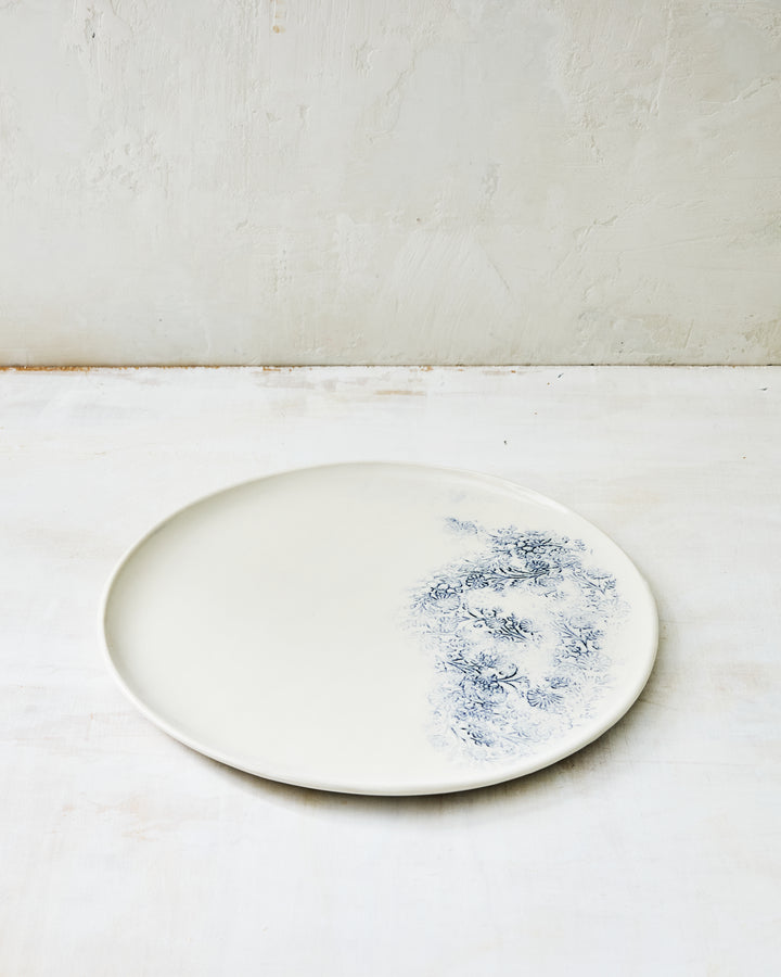 Handmade Kashmir dinner plate in white porcelain with a soft blue indigo floral motif along one edge, shown on a light textured surface.