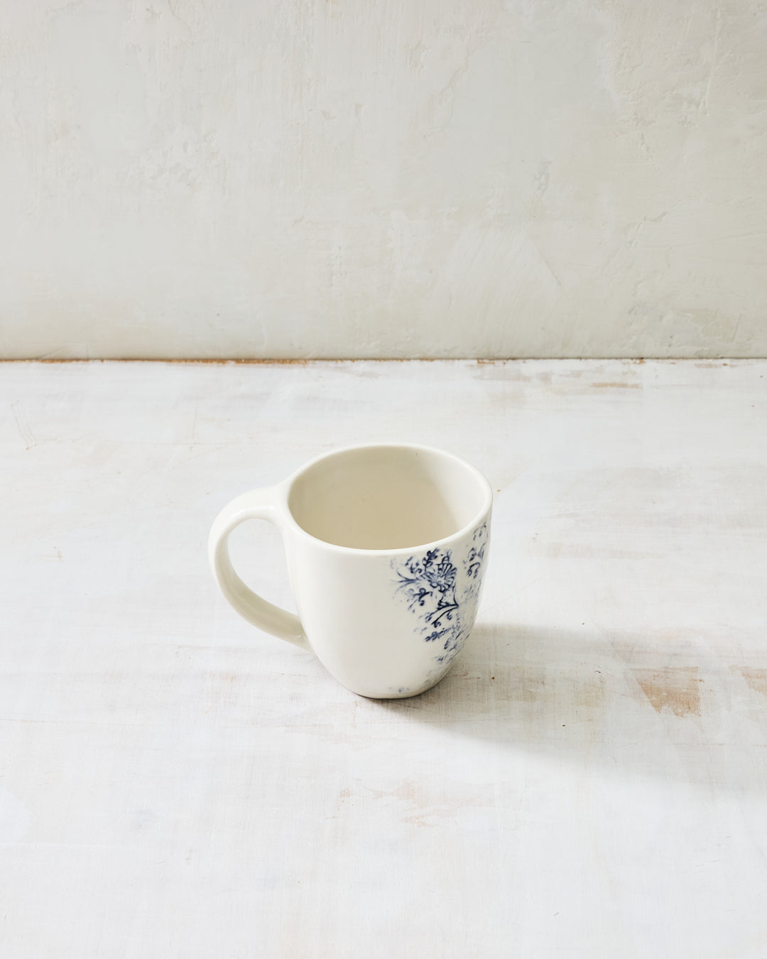Kashmir ceramic mug in glazed white porcelain with hand-applied indigo floral pattern, shown on a light plaster surface.