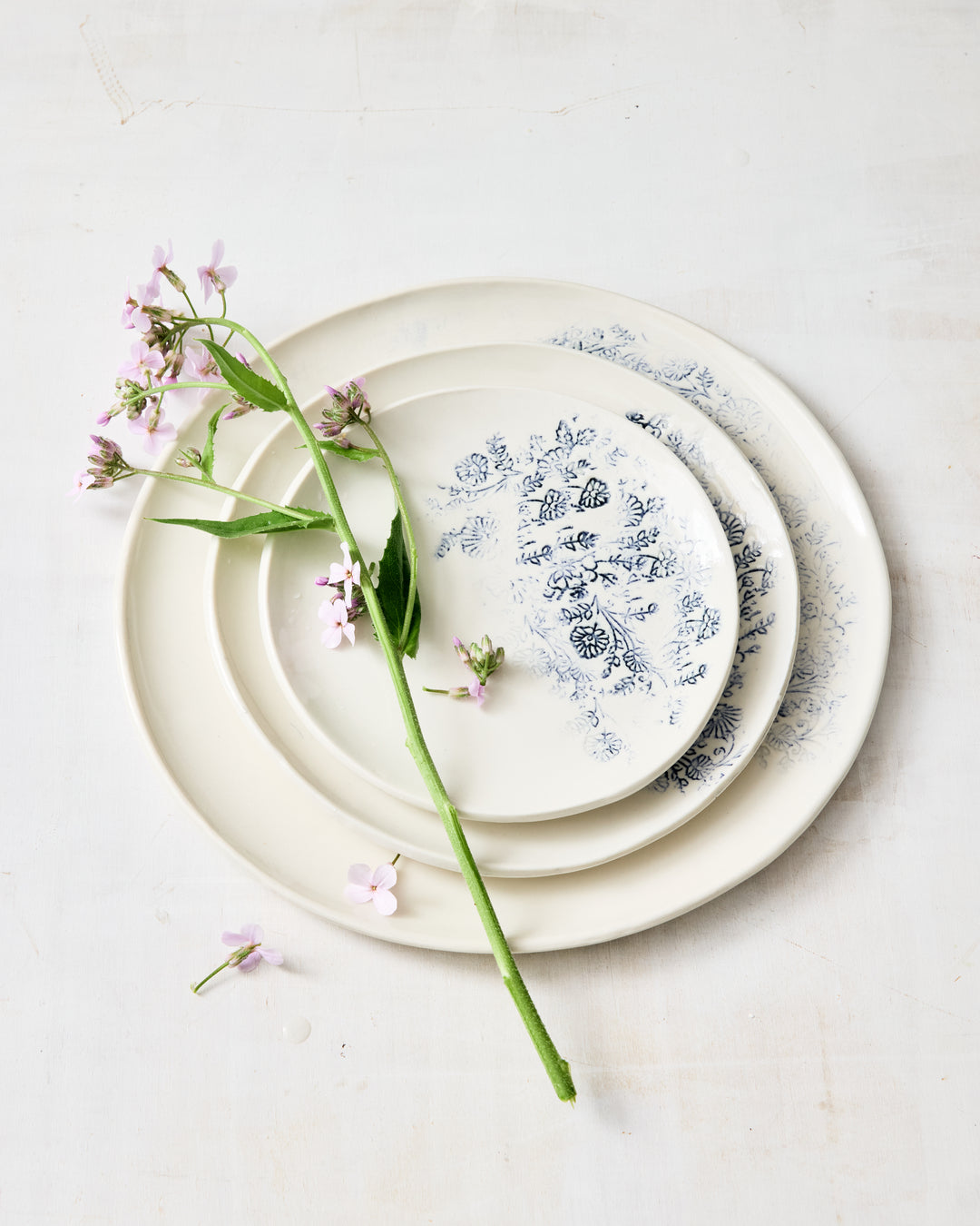 Stack of Kashmir porcelain plates in varying sizes, finished in white with indigo blue floral motifs, styled with fresh pink flowers on a light studio surface.