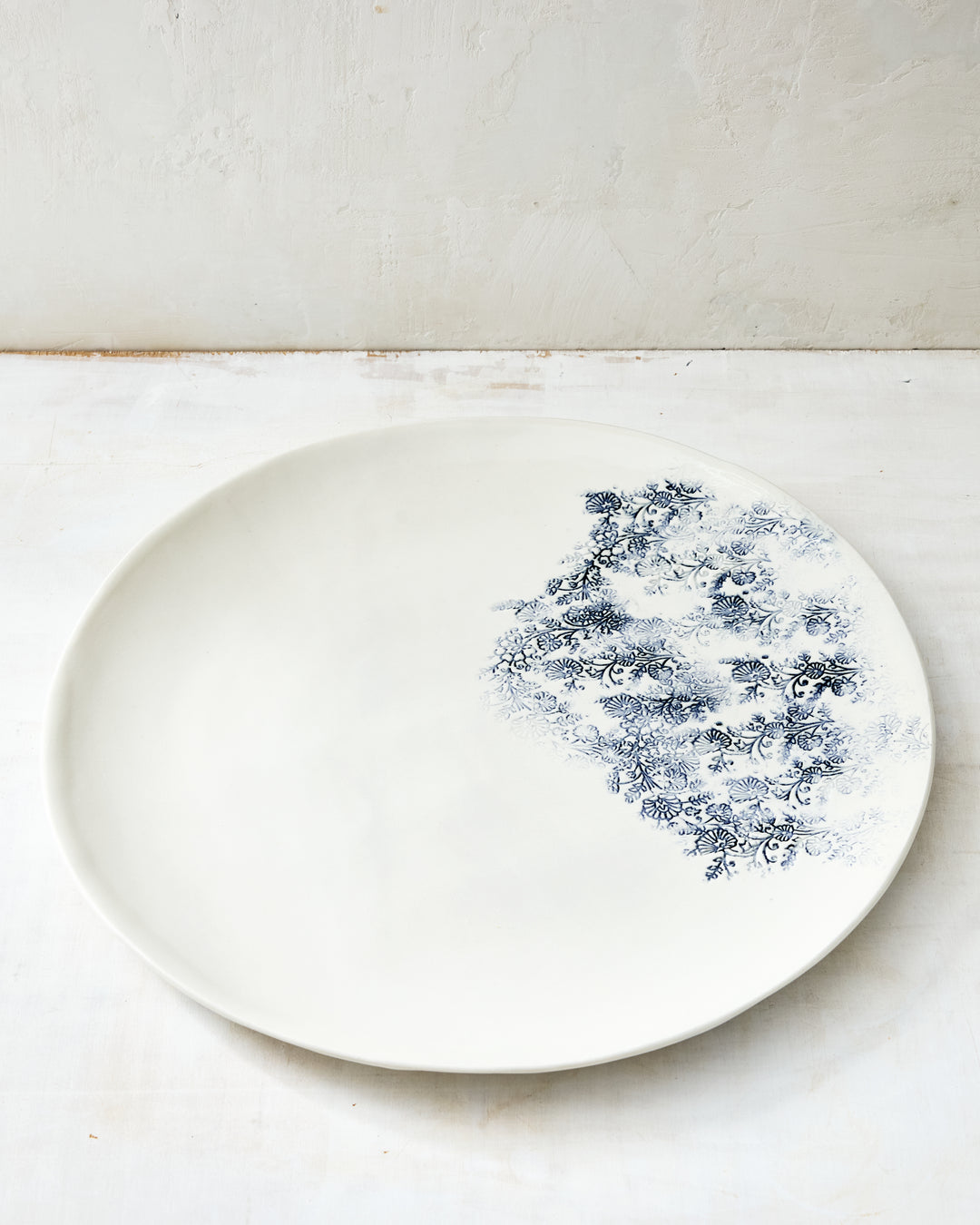 Handmade Kashmir round tray in white ceramic stoneware, featuring a soft blue floral transfer pattern inspired by antique textiles, with a wide, gently curved rim and minimalist form, photographed on a light, textured studio surface.