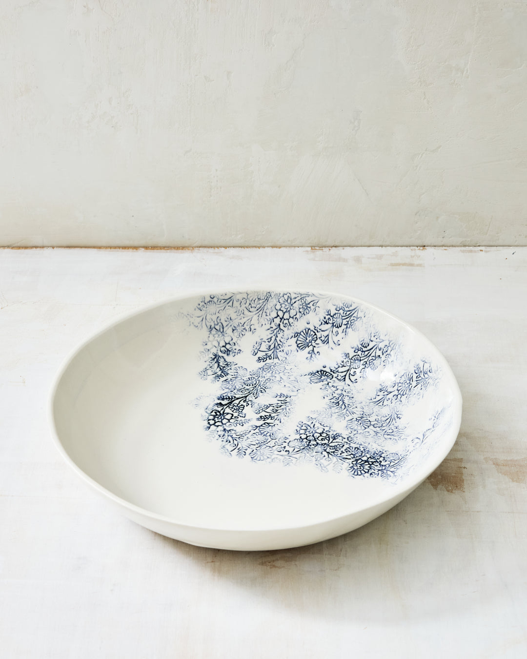 Handcrafted Kashmir serving bowl in white ceramic stoneware, featuring a soft blue floral transfer pattern inspired by antique textiles, with a wide, gently sloped profile ideal for serving salads, pasta, or shared dishes, photographed on a light, textured studio surface.