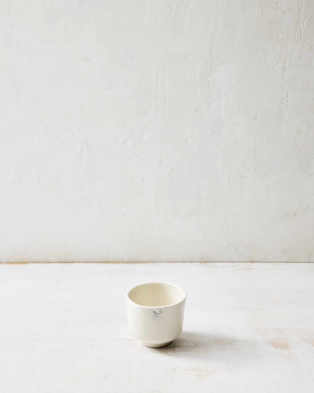 Kashmir tea bowl in glazed porcelain with subtle indigo floral detail, shown on a white plaster surface.