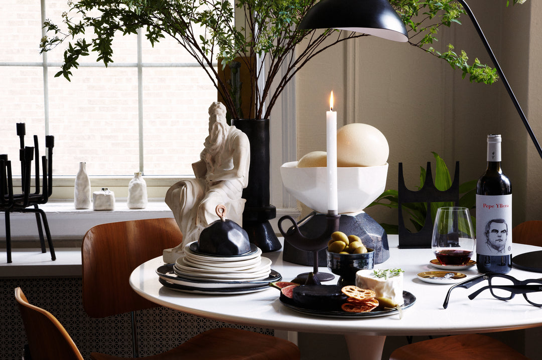 DBO HOME artisan ceramic plates and blows stacked on elegant apartment table with appetizers, a bottle of wine, a candle and a small statue in front of a large window and houseplant | Photo by Marcus Hay, styling by Hallie Burton