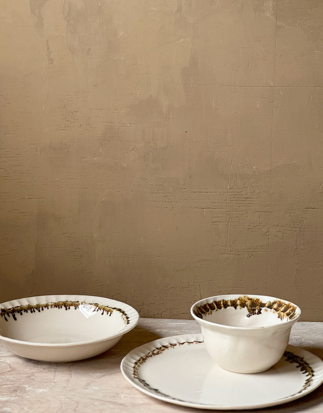 Three distinctive coordinating ceramic pieces from the DBO HOME Nonna Collection - a dinner plate, pasta bowl, and small bowl - sit on a wooden surface against a caramel colored wooden backdrop. Each piece is an off-white natural white with a delicate and distinctive fluted rim, adorned with organic and unique bronze glaze drips and splashes in a delicate ring at the base of the rim.