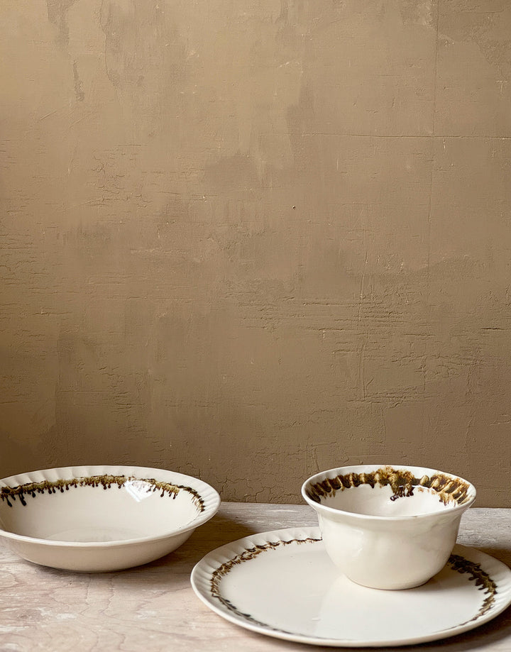 Three distinctive coordinating ceramic pieces from the DBO HOME Nonna Collection - a dinner plate, pasta bowl, and small bowl - sit on a wooden surface against a caramel colored wooden backdrop. Each piece is an off-white natural white with a delicate and distinctive fluted rim, adorned with organic and unique bronze glaze drips and splashes in a delicate ring at the base of the rim.