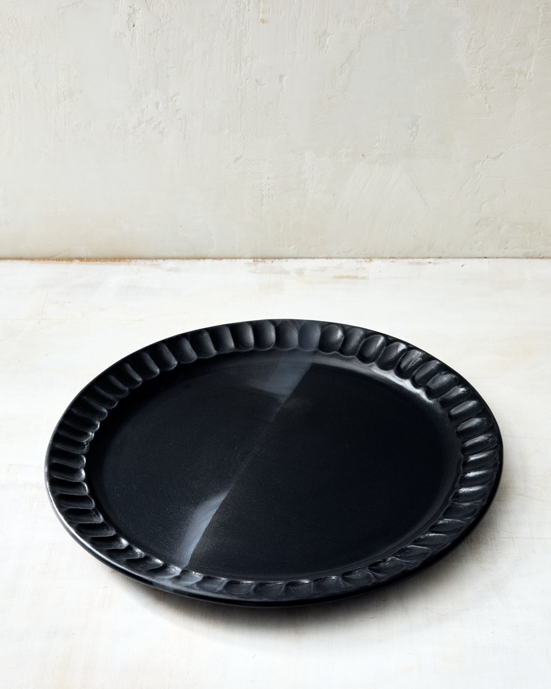 DBO HOME Nonna Dinner Plate in matte black porcelain, featuring a softly scalloped rim and natural undulations that show its handmade origin. The deep, satin-black glaze has a velvety finish that absorbs light. Shown against a pale rustic surface for contrast.