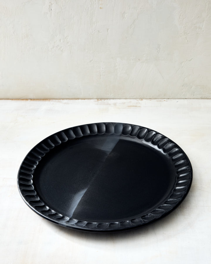 DBO HOME Nonna Dinner Plate in matte black porcelain, featuring a softly scalloped rim and natural undulations that show its handmade origin. The deep, satin-black glaze has a velvety finish that absorbs light. Shown against a pale rustic surface for contrast.