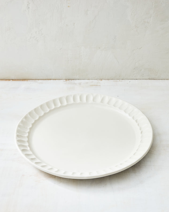 DBO HOME Nonna Dinner Plate in porcelain with a shimmering white satin glaze, revealing the natural warmth of the clay beneath. The softly fluted rim and gentle curves echo vintage European forms, while remaining modern and minimal. Photographed on a neutral-toned surface in soft, natural light.