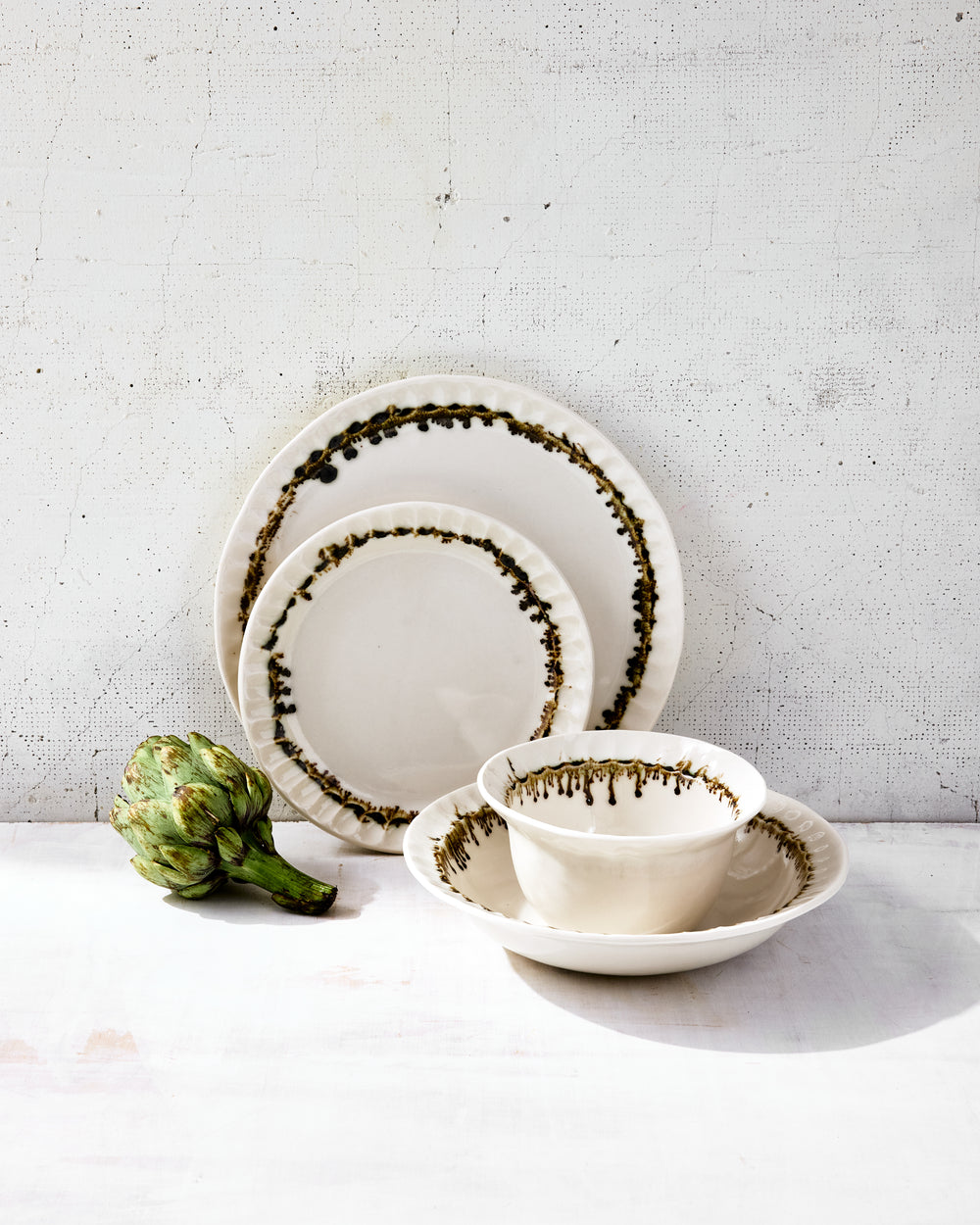 DBO HOME Nonna Dinnerware Set in clear-glazed porcelain with hand-painted bronze rims, styled with a fresh green artichoke. Shown are a dinner plate, salad plate, wide bowl, and small bowl, all featuring softly fluted edges and subtle asymmetry that reflect the handmade process. The scene is warm, natural, and quietly elevated.
