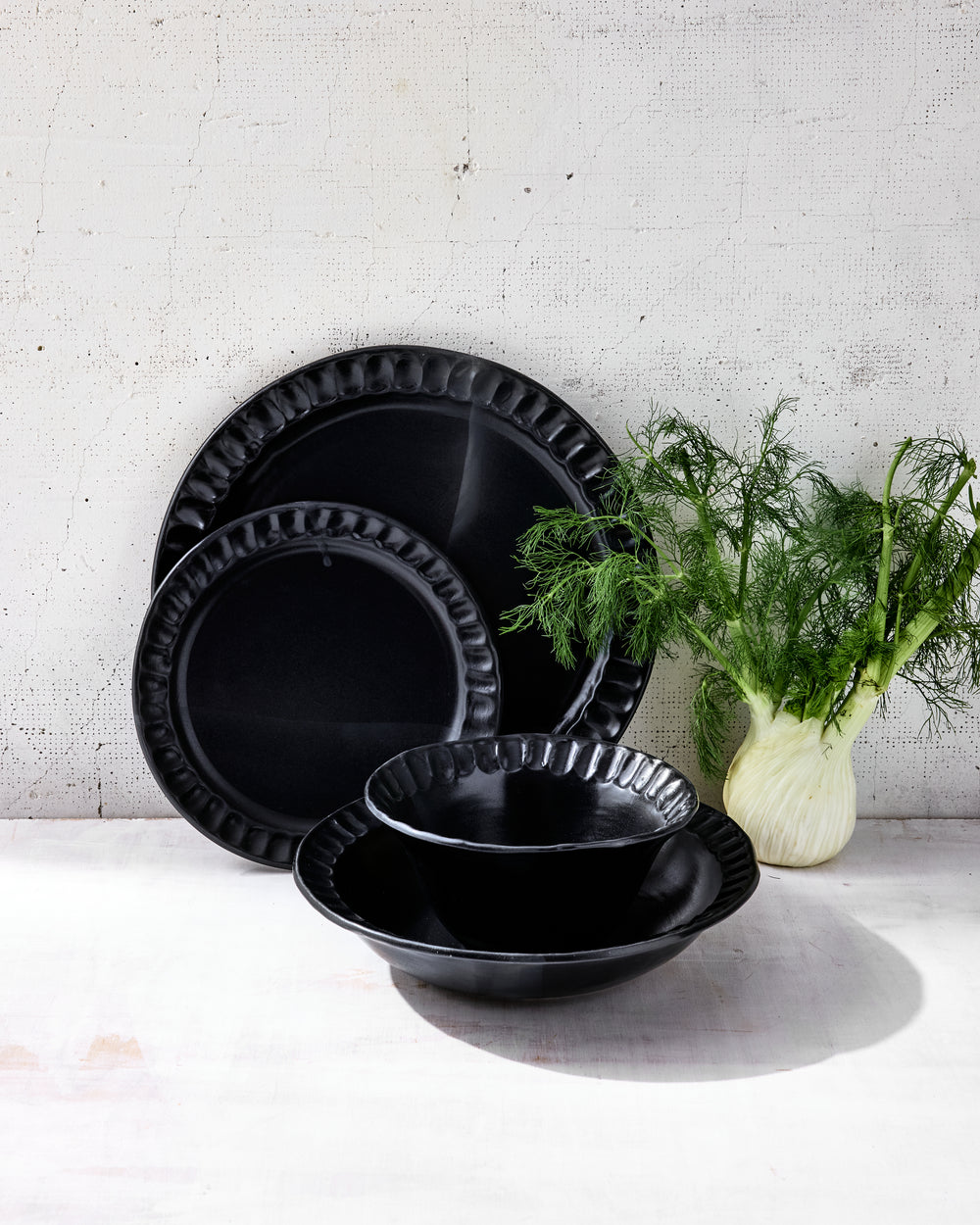 DBO HOME Nonna Dinnerware in Matte Black glazed porcelain, styled with fresh fennel on a pale surface. Shown are softly fluted plates and bowls with a satin glaze that creates rich depth and a slightly reflective finish. A grounded, minimal composition that speaks to quiet luxury.