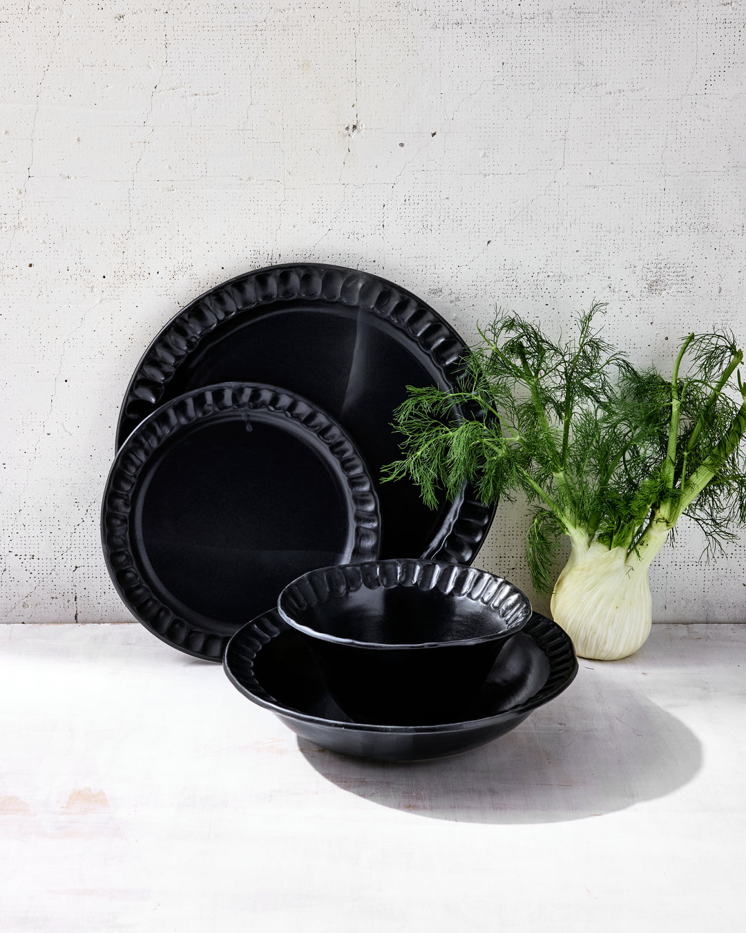 DBO HOME Nonna Dinnerware in Matte Black glazed porcelain, styled with fresh fennel on a pale surface. Shown are softly fluted plates and bowls with a satin glaze that creates rich depth and a slightly reflective finish. A grounded, minimal composition that speaks to quiet luxury.