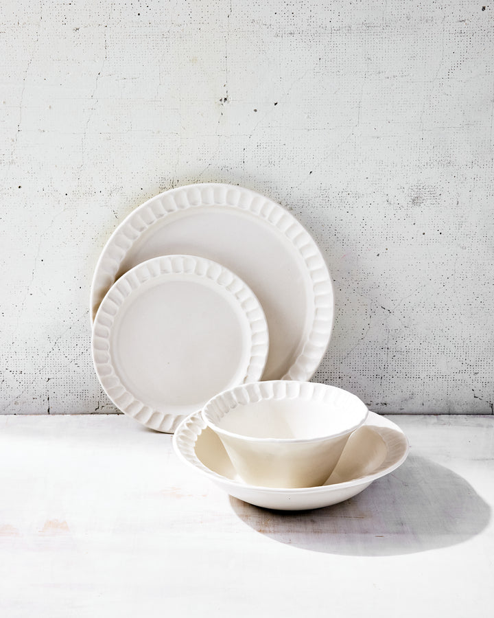 Nonna Dinnerware in shimmering Snowflake-glazed porcelain by DBO HOME, stacked to show subtle rim variations and soft undulations across the plates and bowls. The finish is warm, matte, and tactile.