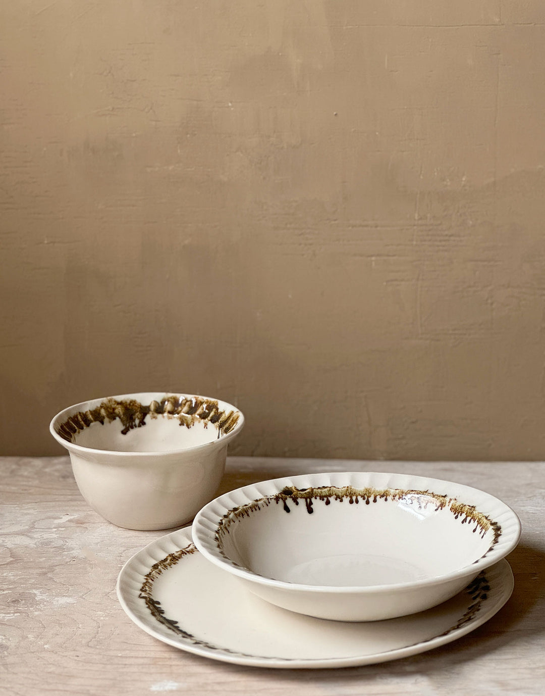 Three distinctive coordinating ceramic pieces from the DBO HOME Nonna Collection - a dinner plate, pasta bowl, and small bowl - sit on a wooden surface against a caramel colored wooden backdrop. Each piece is an off-white natural white with a delicate and distinctive fluted rim, adorned with organic and unique bronze glaze drips and splashes in a delicate ring at the base of the rim.