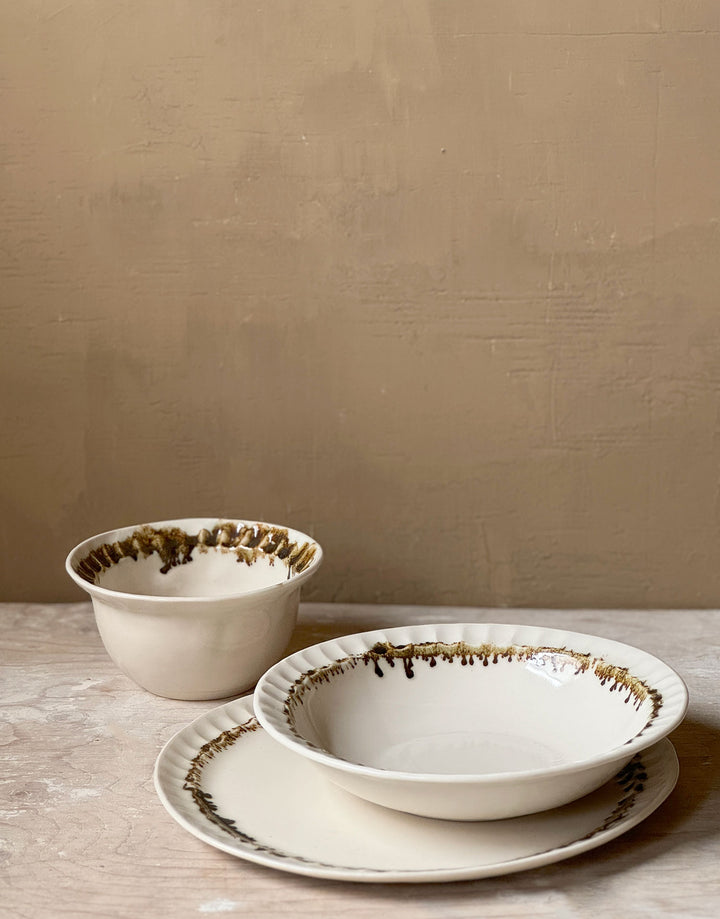 Three distinctive coordinating ceramic pieces from the DBO HOME Nonna Collection - a dinner plate, pasta bowl, and small bowl - sit on a wooden surface against a caramel colored wooden backdrop. Each piece is an off-white natural white with a delicate and distinctive fluted rim, adorned with organic and unique bronze glaze drips and splashes in a delicate ring at the base of the rim.