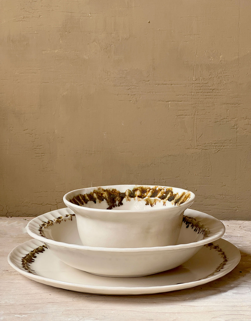 Three distinctive coordinating ceramic pieces from the DBO HOME Nonna Collection - a dinner plate, pasta bowl, and small bowl - sit nested on a wooden surface against a caramel colored wooden backdrop. Each piece is an off-white natural white with a delicate and distinctive fluted rim, adorned with organic and unique bronze glaze drips and splashes in a delicate ring at the base of the rim.