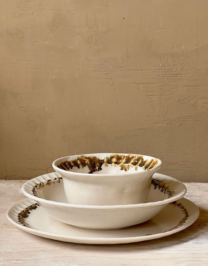 Three distinctive coordinating ceramic pieces from the DBO HOME Nonna Collection - a dinner plate, pasta bowl, and small bowl - sit nested on a wooden surface against a caramel colored wooden backdrop. Each piece is an off-white natural white with a delicate and distinctive fluted rim, adorned with organic and unique bronze glaze drips and splashes in a delicate ring at the base of the rim.