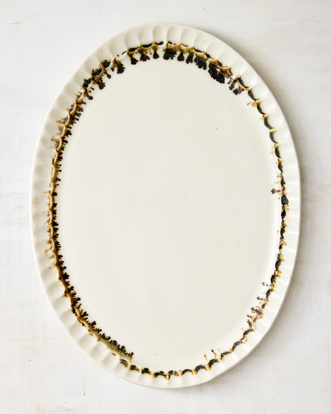 DBO HOME Nonna Oval Platter in Bronze Drip, handmade from white porcelain with a clear glossy glaze and rimmed in hand-applied bronze that drips like espresso at the scalloped edge, shown flat on a pale textured background