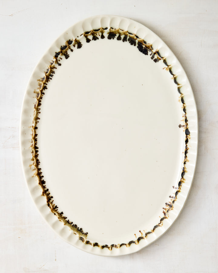 DBO HOME Nonna Oval Platter in Bronze Drip, handmade from white porcelain with a clear glossy glaze and rimmed in hand-applied bronze that drips like espresso at the scalloped edge, shown flat on a pale textured background