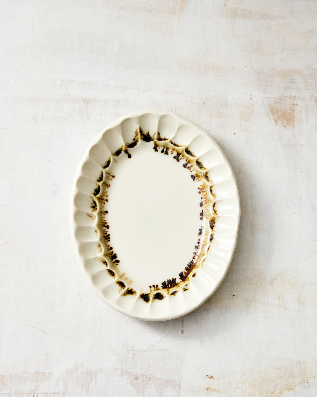 DBO HOME Nonna Oval Side Dish in Bronze glaze, with deeply textured scalloped edges and bronze detailing hand-applied over clear-glazed white porcelain, photographed from above on a soft rustic surface