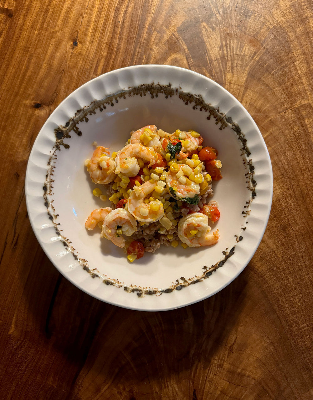 In this closeup of the Nonna Pasta Bowl - a glossy organic off-white ceramic bowl with a shallow belly and a fluted, ruffled edge, adorned with painterly drips and splashes of bronze glaze around the iconic rim -  the bowl sits on a warm wood table and is filled with a heaping scoop of a summer shrimp, corn, and tomato salad.