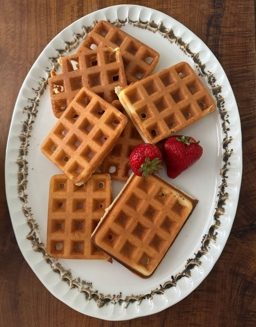 In this closeup of the Nonna Oval Platter - a glossy organic off-white ceramic bowl with a shallow belly and a fluted, ruffled edge, adorned with painterly drips and splashes of bronze glaze around the iconic rim -  the plate sits on a warm wood table and is piled high with small fluffy waffles and two ripe garden strawberries.