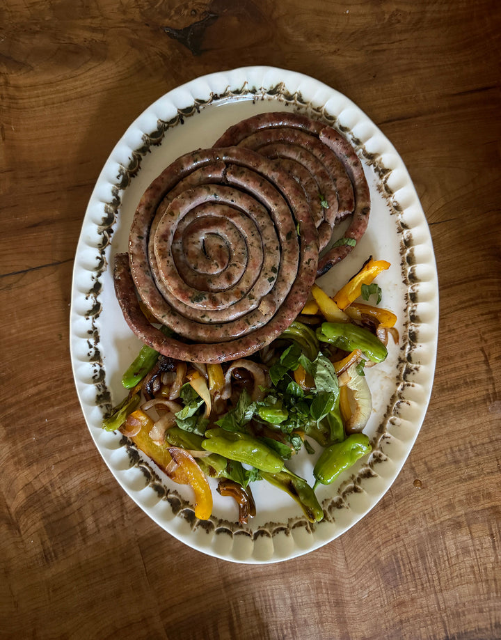 In this closeup of the Nonna Oval Platter - a glossy organic off-white ceramic bowl with a shallow belly and a fluted, ruffled edge, adorned with painterly drips and splashes of bronze glaze around the iconic rim -  the plate sits on a warm wood table and is piled with pinwheels of pork sausage and a mix of grilled summer garden vegetables.