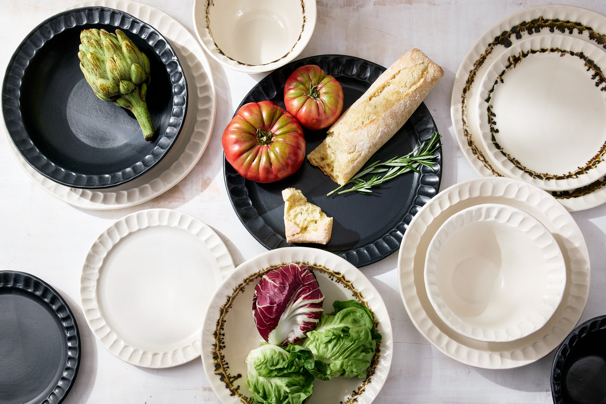 Overhead view of the DBO HOME Nonna dinnerware collection, including dinner plates, salad plates, small bowls, and pasta bowls in Matte Black, Snowflake, and Bronze finishes. Styled with heirloom tomatoes, fresh greens, and rustic bread on a whitewashed surface, highlighting the handmade textures and inviting forms.