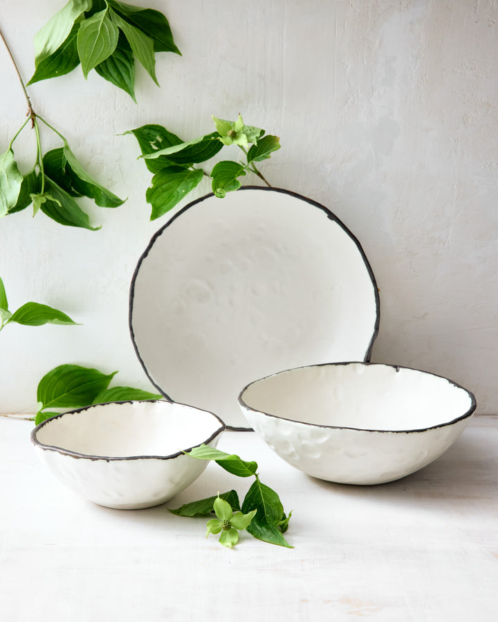 A trio of handcrafted ceramic bowls and plates from DBO Home’s Pinch Collection, displayed with fresh green branches from outside our studio. The creamy white pieces feature softly irregular rims edged in raw dark glaze, showcasing their organic form and dimpled texture. Styled against a soft white backdrop, the composition feels airy, fresh, and quietly elegant.