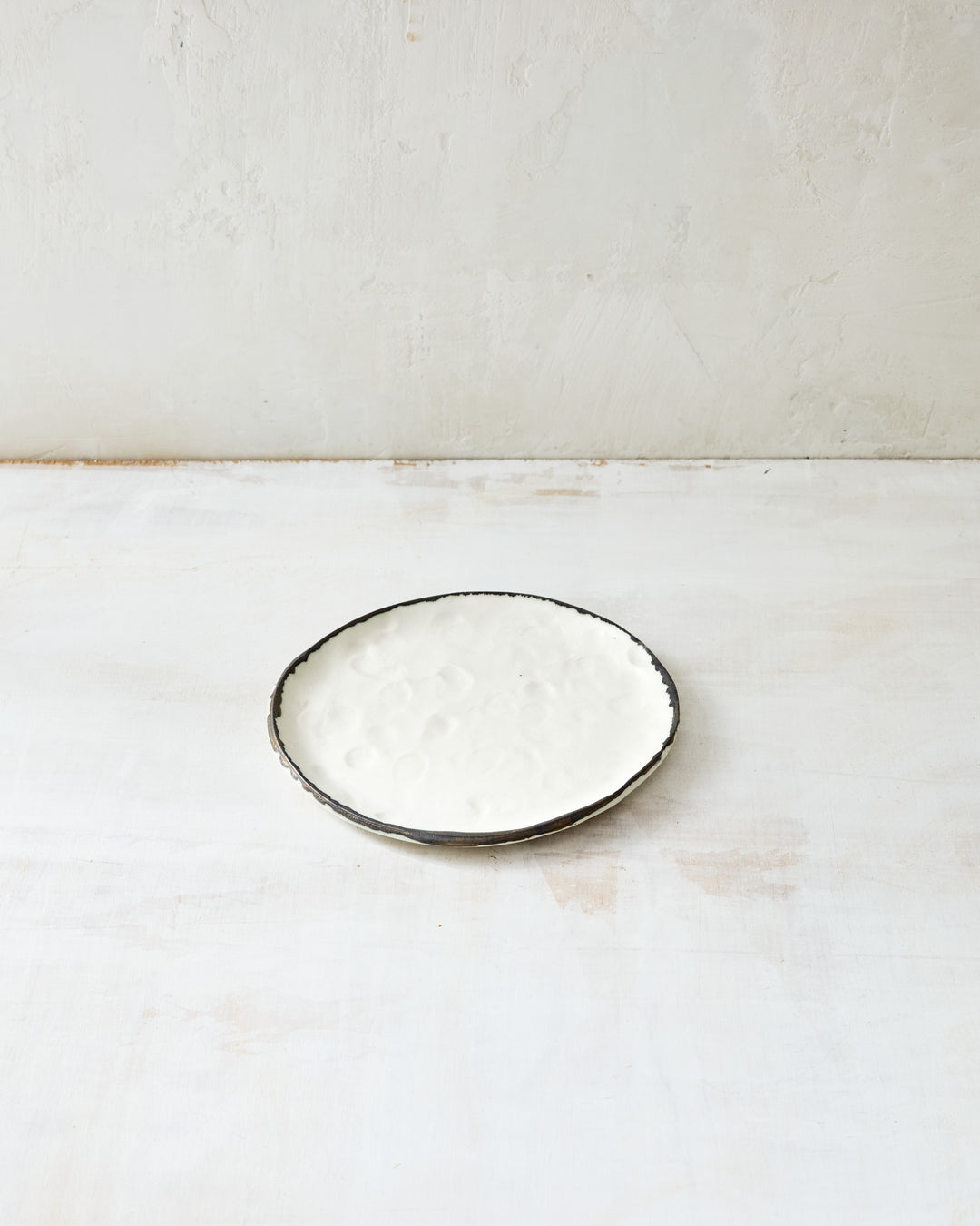 DBO HOME Pinch Bread Plate in creamy white porcelain with a hand-applied bronze rim, shown on a softly distressed white surface. The dimpled texture and irregular edge add quiet character to this small, sculptural plate.