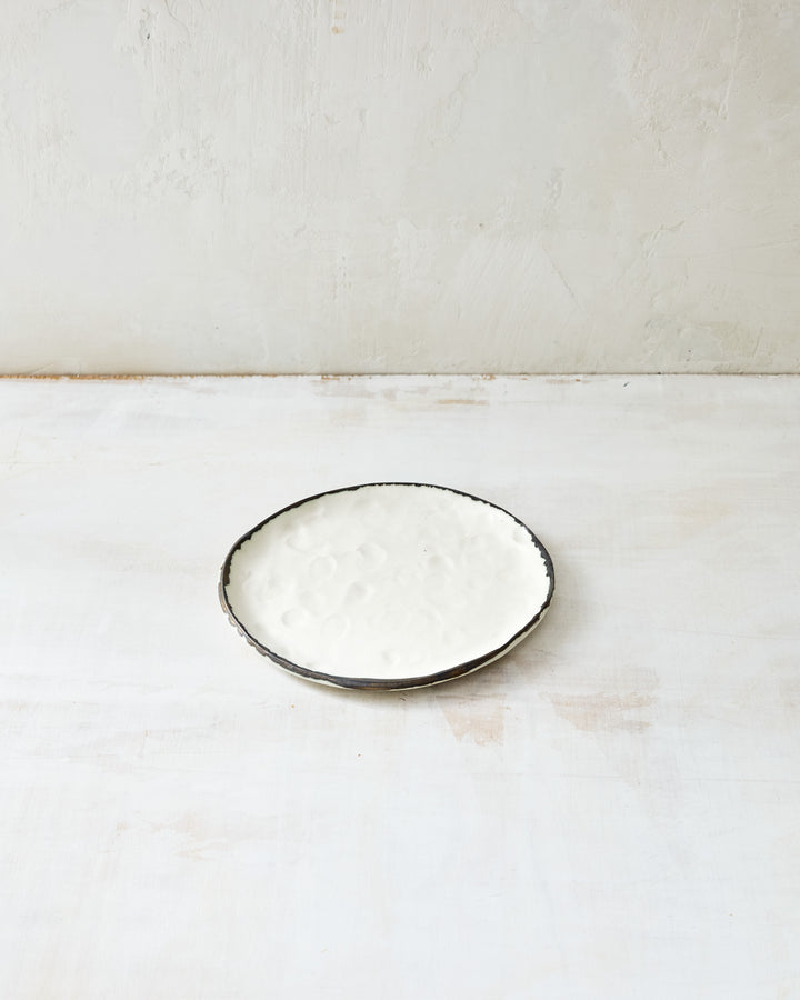 DBO HOME Pinch Bread Plate in creamy white porcelain with a hand-applied bronze rim, shown on a softly distressed white surface. The dimpled texture and irregular edge add quiet character to this small, sculptural plate.
